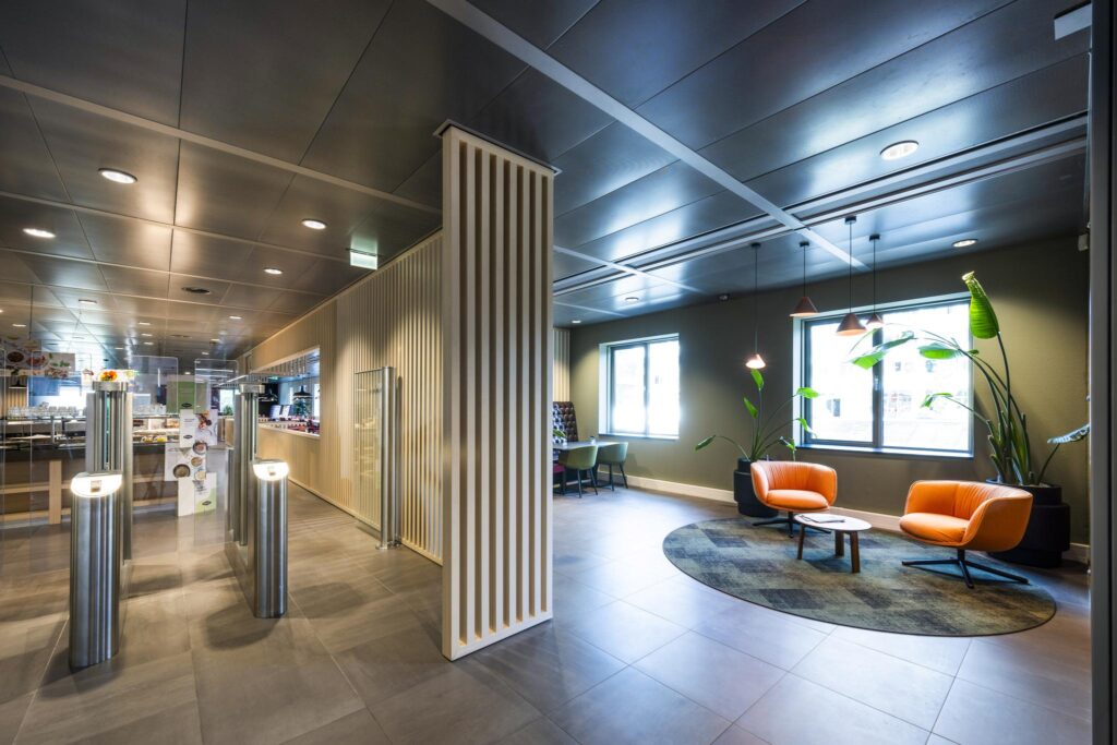Modern lobby area at Koningin Wilhelminaplein with orange lounge chairs, a round rug, and a view into the cafeteria.